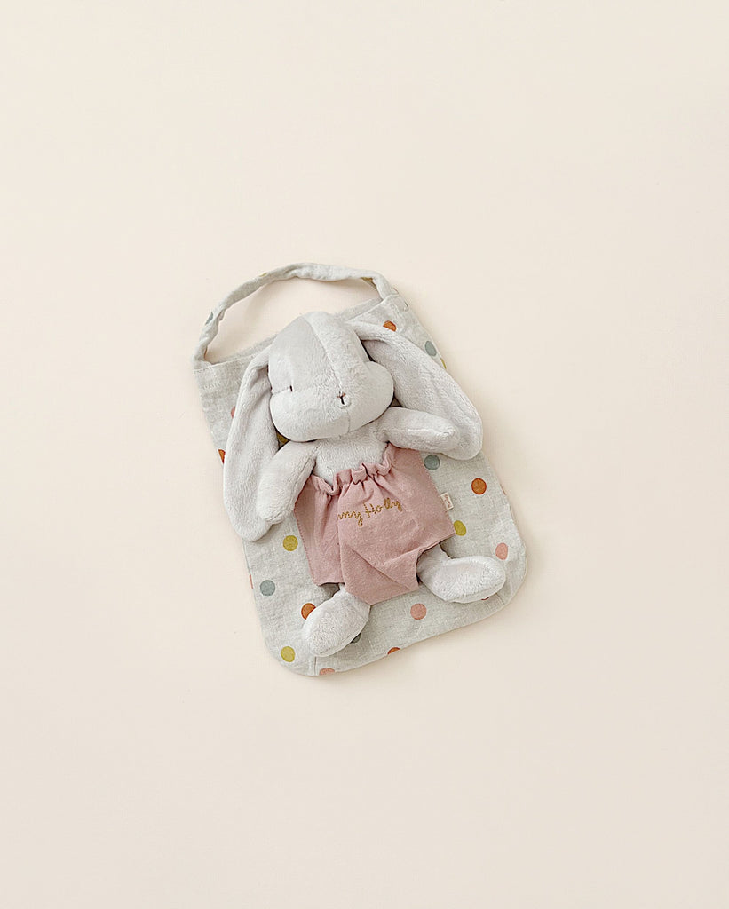 A Maileg Bunny Holly with a pink dress and the name "amy adams" embroidered on it, resting on a pastel polka dot bag against a light beige background.