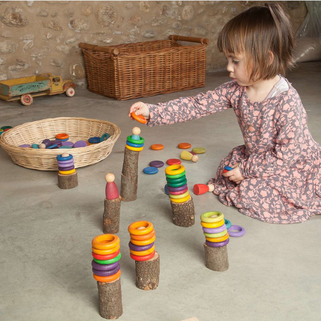 A young child plays with Grapat 36 rings in 6 colors stacked on pegs, sitting on a concrete floor surrounded by a wicker basket and a wooden toy truck.