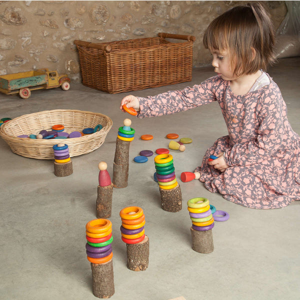 A young child plays with Grapat 36 rings in 6 colors stacked on pegs, sitting on a concrete floor surrounded by a wicker basket and a wooden toy truck.