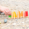 A child's hand setting up colorful Grapat Sticks Gnomes wooden toy figures on a gravel surface, with one toy isolated from a line of others resembling a game setup.