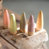 Five Grapat Sticks Gnomes painted in soft pastel colors of pink, green, blue, gray, and gold, standing on a rustic wooden surface, each designed as open-ended toys with simple dotted eyes.