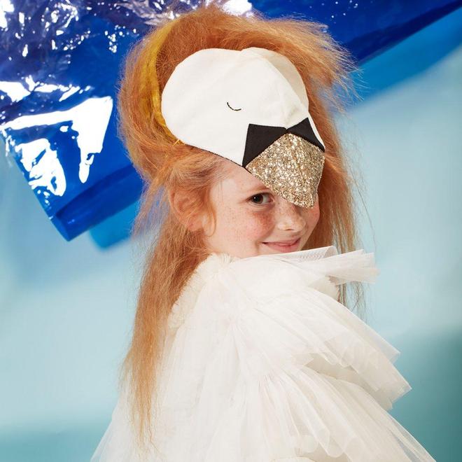 A young girl in a Meri Meri Swan Costume - Final Sale smiles at the camera. She wears a white ruffled outfit and a whimsical mask resembling a bird's face, with gold and black accents, complemented by t