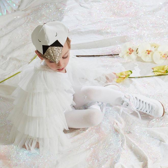 A child in a Meri Meri Swan Costume, wearing sneakers and a velvet headdress, sitting among glittering fabric and scattered flowers.
