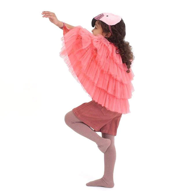 A young girl in a Meri Meri Flamingo Costume playfully poses on one leg, with one arm extended, against a white background.