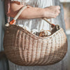 A woman in a light dress holds a large natural rattan Basket of Lavender Felt Flowers filled with fresh vegetables, illuminated by sunlight.
