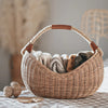 A Basket of Lavender Felt Flowers with a sturdy handle, filled with neatly folded towels in shades of white and gray, sits on a soft, textured surface with a blurred background that includes a decor accent.