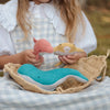 A young girl engages in imaginative play with a basket, holding an Olli Ella Holdie Folk Felt Ocean Animals plush toy and playing with a blue whale and a yellow shark toy, donned in a white lace blouse