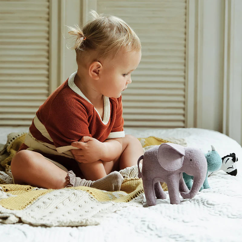A toddler with a ponytail, dressed in a red and white outfit, sits on a knitted blanket, engaging in imaginative play with Olli Ella Holdie Folk Felt Safari Animals including a small stuffed elephant.