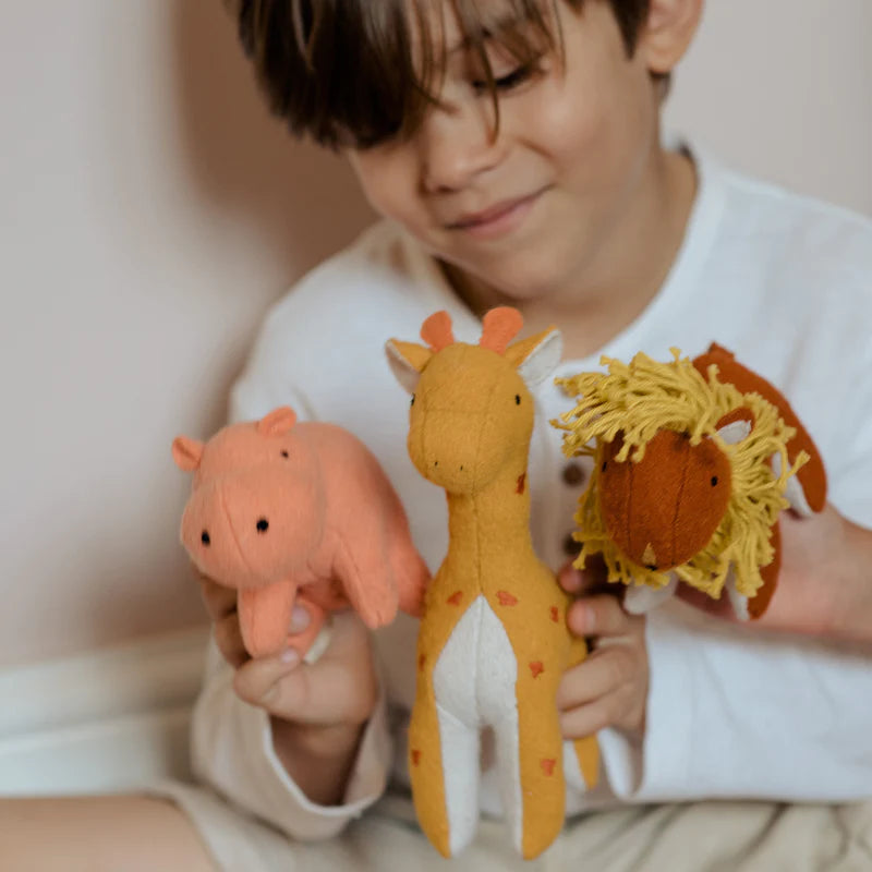 A young boy smiling as he holds up three Olli Ella Holdie Folk Felt Savannah Animals: a pig, a giraffe, and a lion, clearly delighted by his wildlife toys collection.