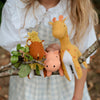 A person holding a branch adorned with three Olli Ella Holdie Folk Felt Savannah Animals: a lion with a mane, a pig, and a giraffe, amidst a natural outdoor setting.