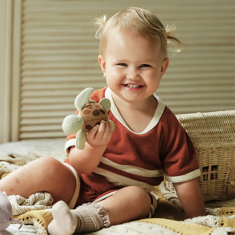 A joyful toddler with light hair in pigtails, wearing a red and white outfit, holds a plush turtle toy from Olli Ella Holdie Folk Felt Marine Animals while smiling broadly, sitting on a cozy blanket indoors.