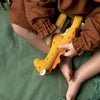 A toddler in a brown outfit holding a Olli Ella Holdie Folk Felt Savannah Animals yellow stuffed dinosaur toy with red spots, sitting on a green blanket. Only the lower half of the child’s body and hands are visible.
