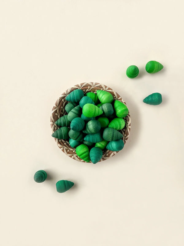 A woven basket filled with green and teal colored shell-shaped beads on a light beige background, designed as Grapat Mandala Trees components, with a few beads scattered around the basket.