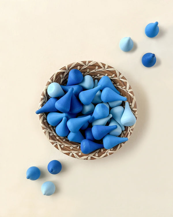 A round wicker basket filled with various shades of blue pebbles, some shaped like hearts from Grapat Mandala Raindrops, on a light beige background. A few pebbles are scattered outside