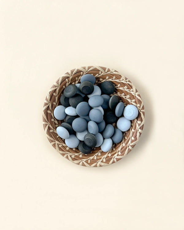 A round woven basket filled with a collection of smooth, grey and blue Grapat Mandala Stones of various shades, arranged neatly on a light beige background.
