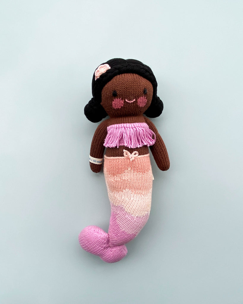 A Cuddle + Kind Maya the Mermaid doll with dark skin, adorned in a pink and white tail, a lilac top, and black hair with a pink flower, against a plain grey background.