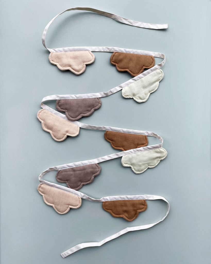 An array of eight Cloud Garland sleep masks, crafted from Oeko-tex certified organic cotton, in various shades like white, beige, grey, and brown, shaped like clouds with attached white ribbons.