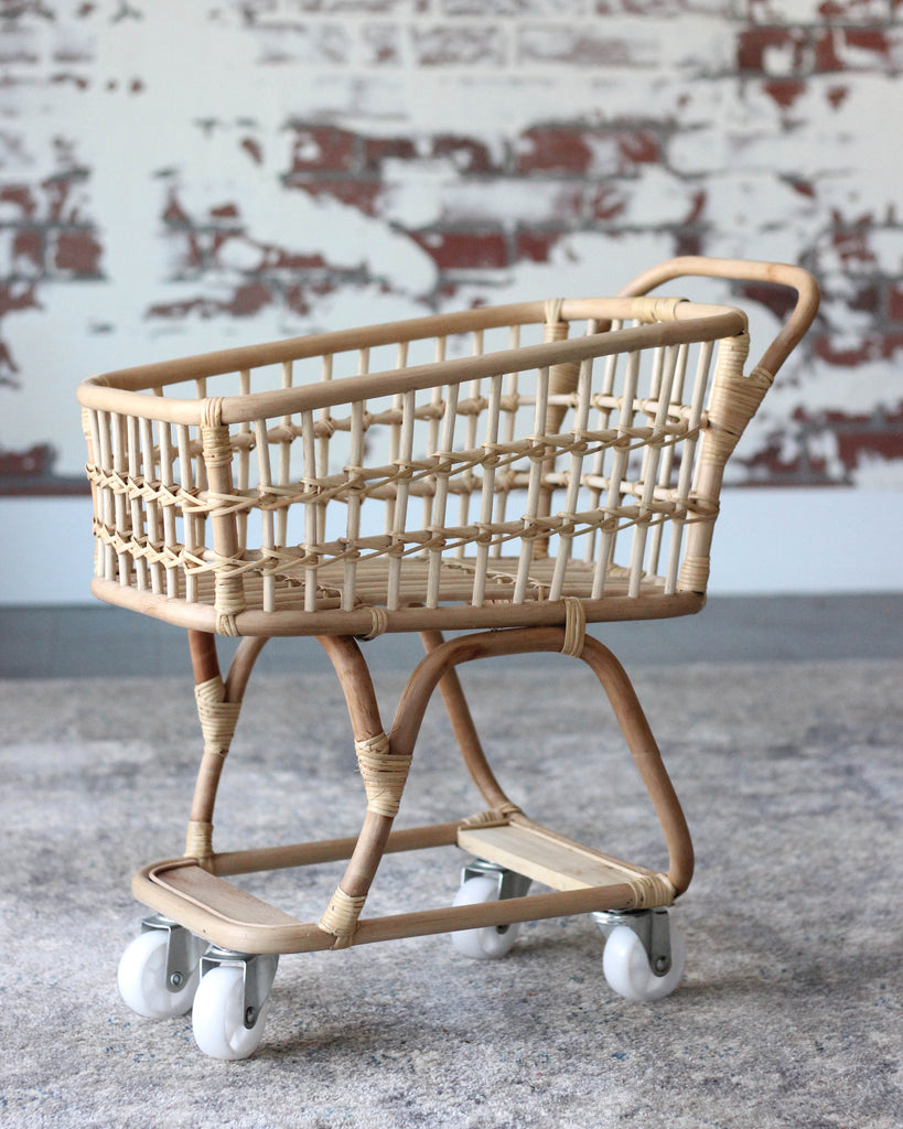 A vintage Rattan Grocery Shopping Cart with large white wheels, set against a wall with a faded brick motif. The shopping cart has a sturdy cane handle and a spacious design.