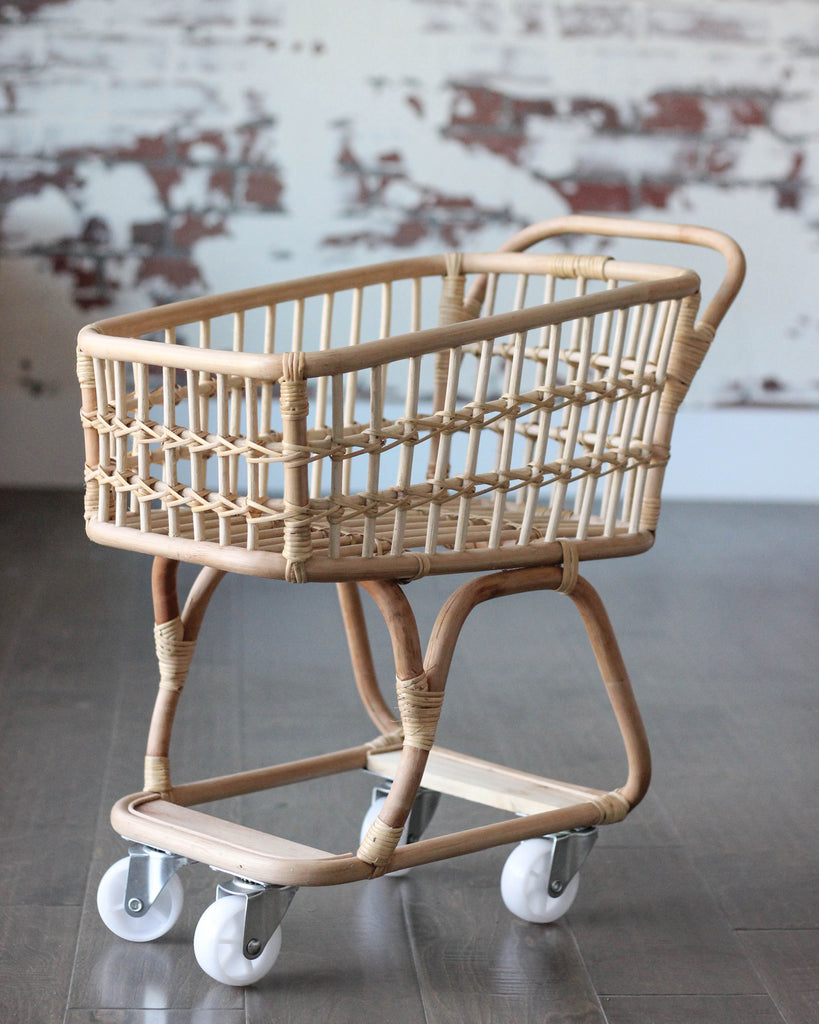 A wooden baby carriage with wicker basket design and four white wheels, displayed in a room with a wall decorated by a mural depicting faded red figures, transforms into a Rattan Grocery Shopping Cart perfect for imaginative pretend play.