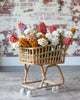 A Rattan Grocery Shopping Cart filled with colorful knitted flowers, including shades of red, orange, white, and purple, standing against a rustic brick wall background.