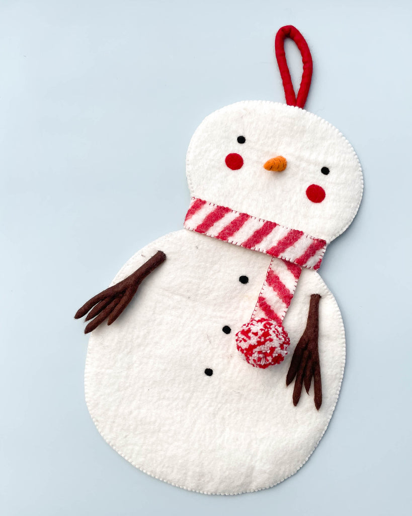 A white organic felt Handmade Snowman Christmas Stocking with a red and white striped scarf, brown twig arms, an orange carrot nose, and three black button details on a light blue background.