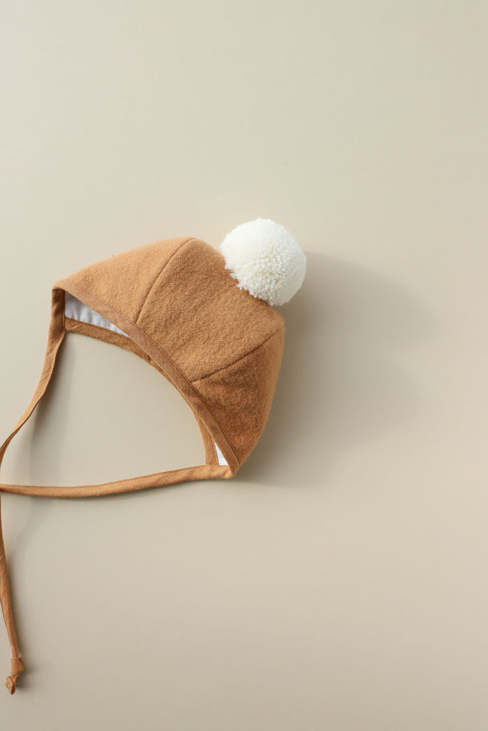 A brown wool Briar Baby Saddle Pom Bonnet with ear flaps and a white pom-pom, lying flat against a light beige background.