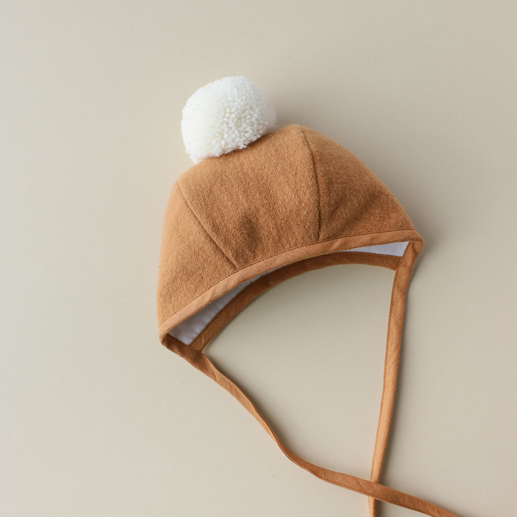 A soft brown Briar Baby Saddle Pom Bonnet with ear flaps and a white pom-pom on top, laid flat on a light beige background.