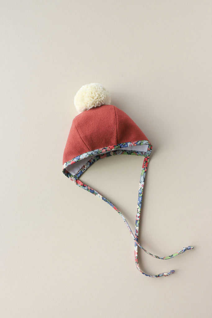 A Briar Baby Blooming Pom Bonnet with a fluffy white pom-pom on top and Liberty of London floral lining, laid flat on a light beige background. The hat has ear flaps and string ties.