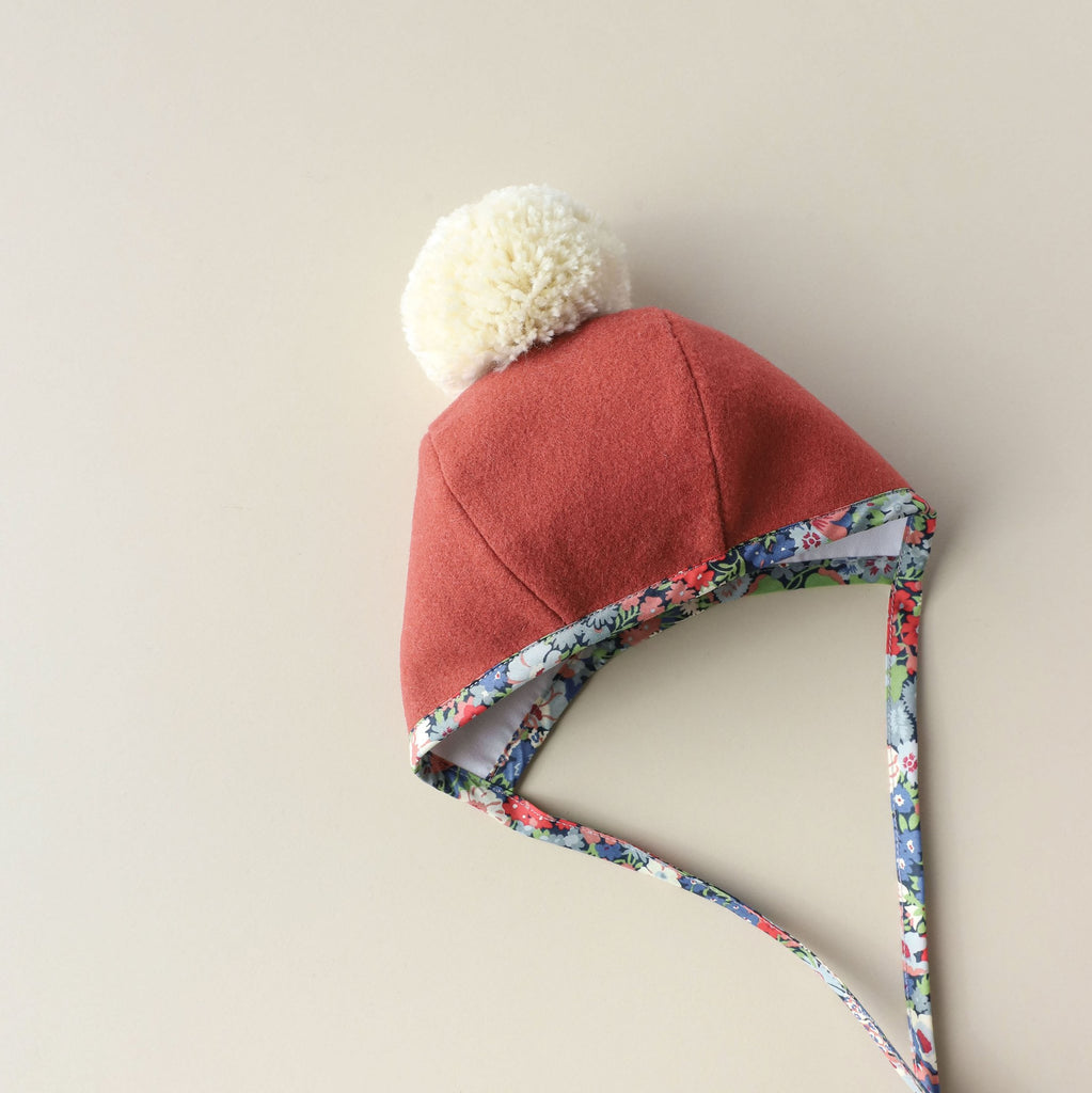 A Briar Baby Blooming Pom Bonnet handmade in the USA, with floral trim and a white pom-pom on top, set against a neutral beige background.