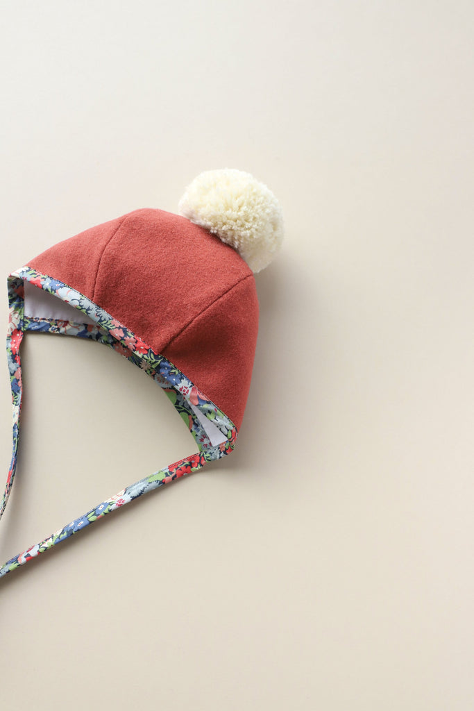 A Briar Baby Blooming Pom Bonnet with a Liberty of London floral print tie, laid flat on a soft beige background.