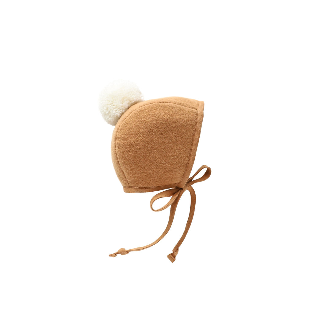 A handmade, Briar Baby Saddle Pom Bonnet with a fluffy white pom-pom on top and thin straps tied into a bow, isolated on a white background.
