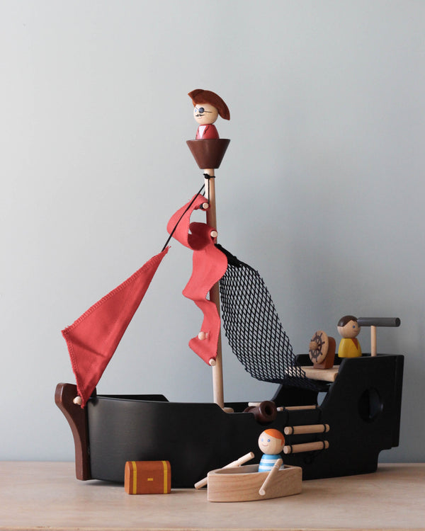 A Pirate Ship Set toy with figurines, featuring a character in a crow's nest, another climbing a rope ladder, and a third by the wheel. The ship has a red flag and detailed components.