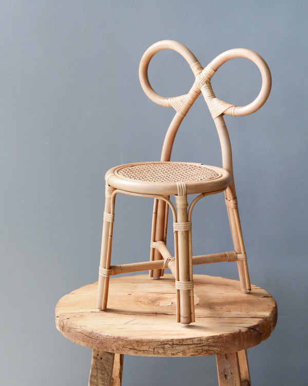 A unique Toddler Rattan Bow Chair made of bamboo, featuring an intricately designed backrest shaped like interlocking loops, stands on a rustic wooden platform against a plain gray background.