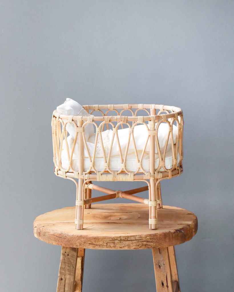 A Poppie Rattan Doll Crib + Duvet Set filled with white pillows, displayed on a rustic wooden stool against a gray wall background.
