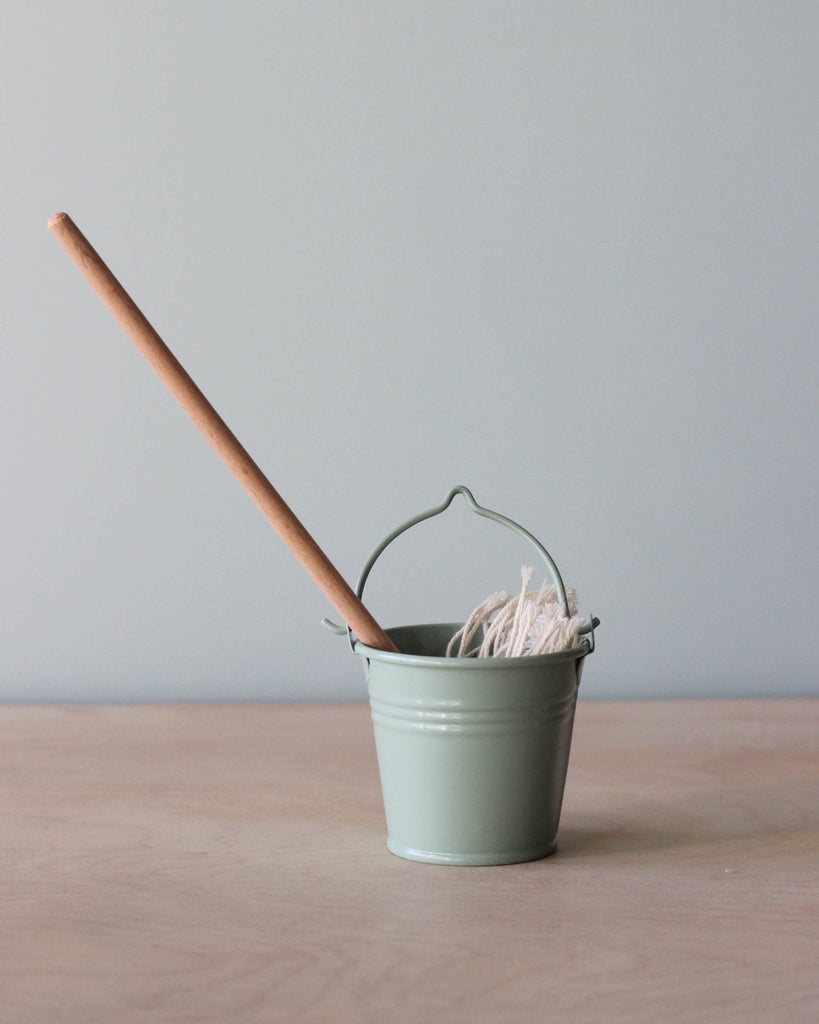 A small light green Maileg Miniature Mop & Bucket with a wooden mop handle, sized perfectly for Mini Maileg friends, containing a white mop head, stands against a soft neutral background.