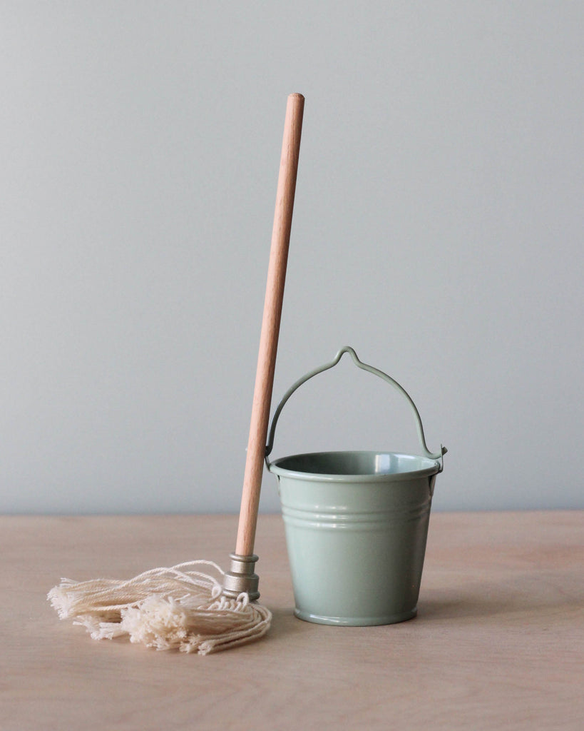 A simple, small green bucket with a metal handle next to a Maileg Miniature Mop & Bucket on a wooden surface against a neutral background, perfect for MINI sized Maileg friends.