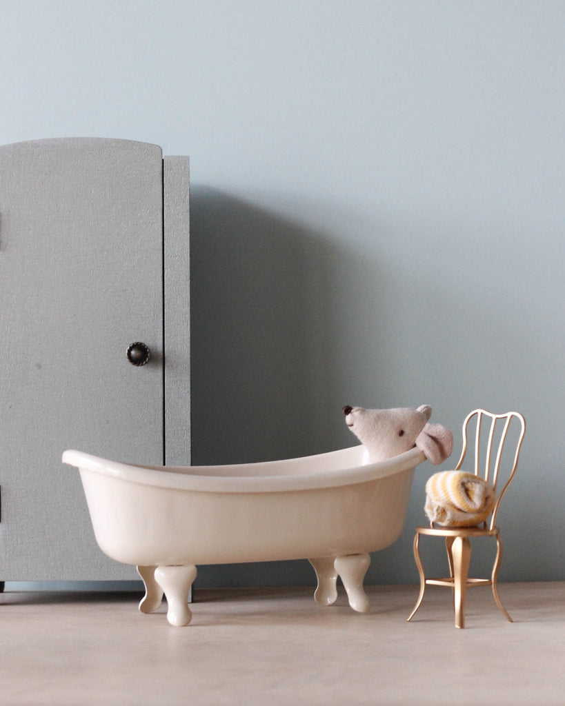 A small stuffed mouse sits in a Maileg Mini Bathtub, facing a toy cupboard and a tiny golden chair with a knitted cushion, all against a soft blue wall.