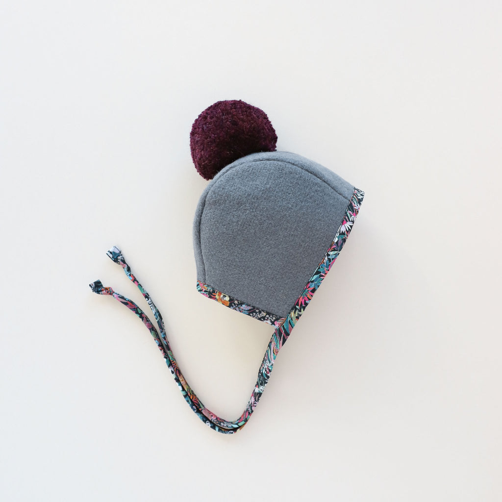 A Briar Baby Florentina Pom Bonnet with a purple pom-pom and floral trim on a plain white background. The bonnet has long, braided ties hanging from each side.