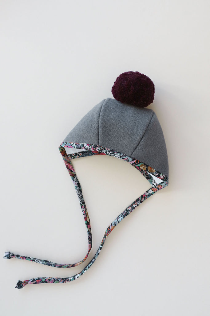 A Briar Baby Florentina Pom Bonnet with a purple pompom on top and multicolored braided ties, lying against a white background.