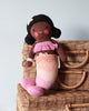 A Cuddle + Kind Maya the Mermaid doll with dark yarn hair and pink clothes sits on a wicker basket against a plain background. The doll sports a smiling face and wears a fringed top and striped pants.