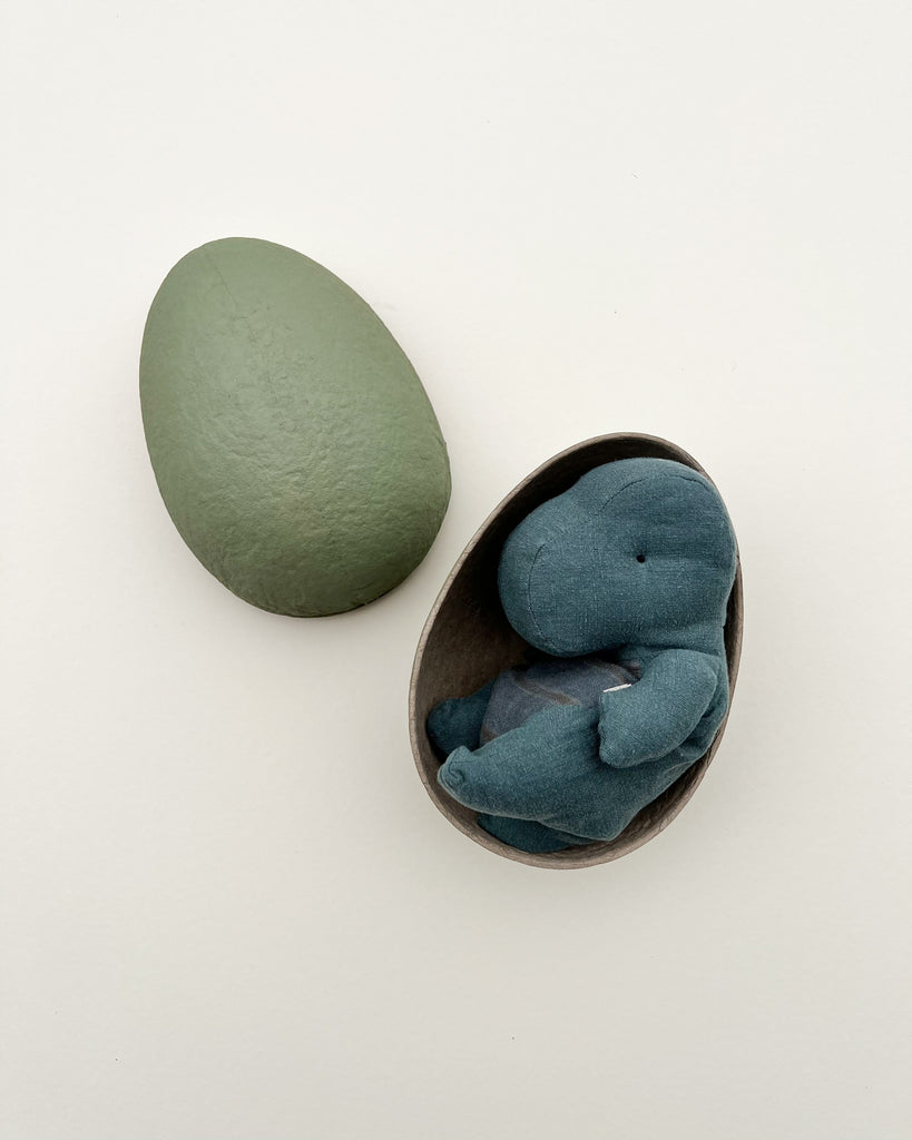 A cuddly blue plush elephant curled up inside a Maileg Small Gantosaurus in egg - Green, with its other half lying next to it. The background is a plain light color.