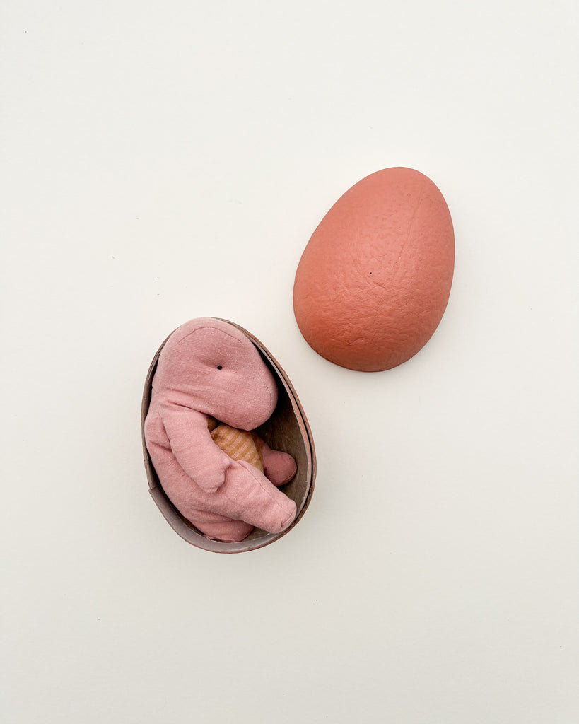 A small Gantosaurus stuffed toy in a pink hue is snugly nestled inside half of a brown, textured egg-shaped container, reminiscent of the delightful surprises from the Maileg collection, with the other half of the container placed nearby on a light beige background.