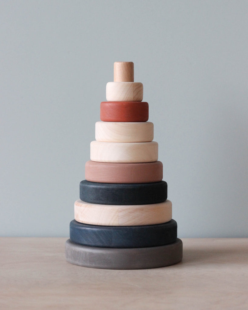 A Wooden Pyramid Stacker - Terracotta consisting of a vertical dowel with stacked rings in various sizes and muted colors, ranging from light to dark, set against a plain, light grey background.