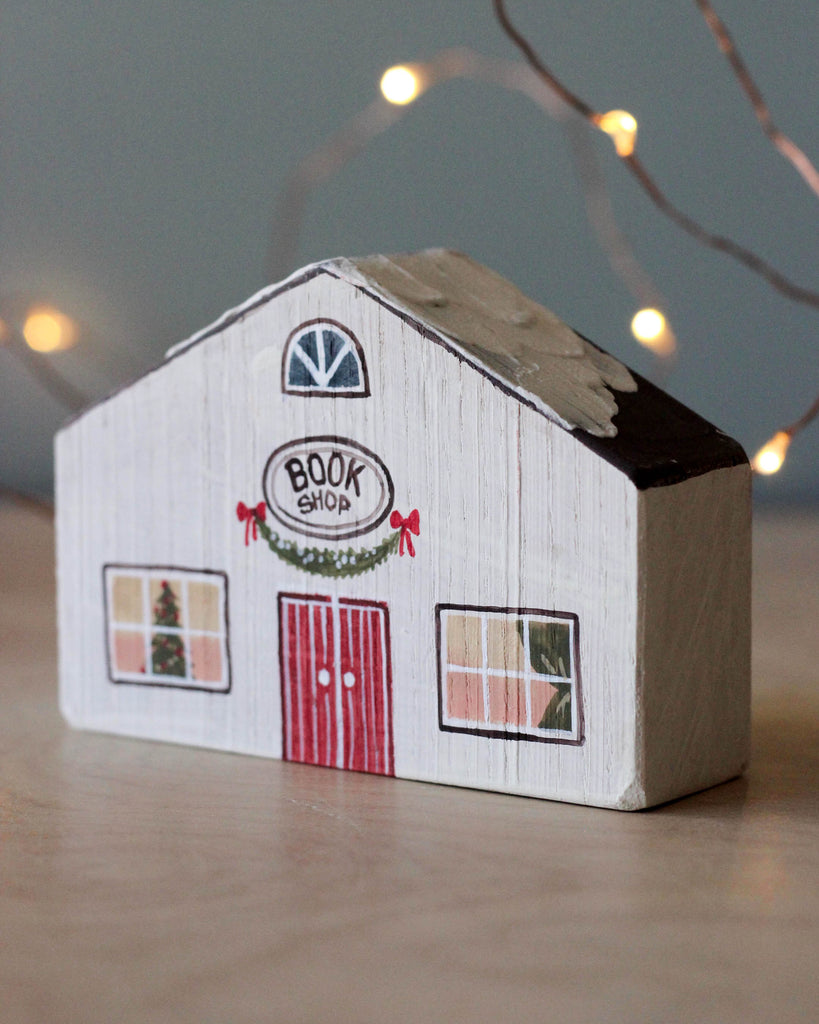 A small, handprinted wooden block painted to resemble a bookshop with a festive holiday theme. The building has a white facade, red double doors, and a wreath above the entrance. The windows show a decorated Christmas tree, perfect for your Handmade Wooden Christmas Village. Soft, warm lights are blurred in the background.