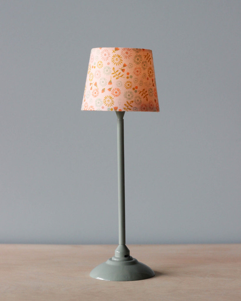 A tall Maileg Miniature Floor Lamp with a grey base and stem, topped with a conical lampshade decorated with orange floral and starburst patterns on a pale background.