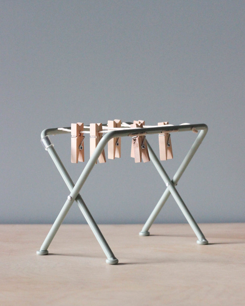A Maileg Miniature Drying Rack with wooden clothespins clipped along its top bars stands on a light wood surface against a plain gray background, resembling a charming dollhouse accessory with its intricate details.
