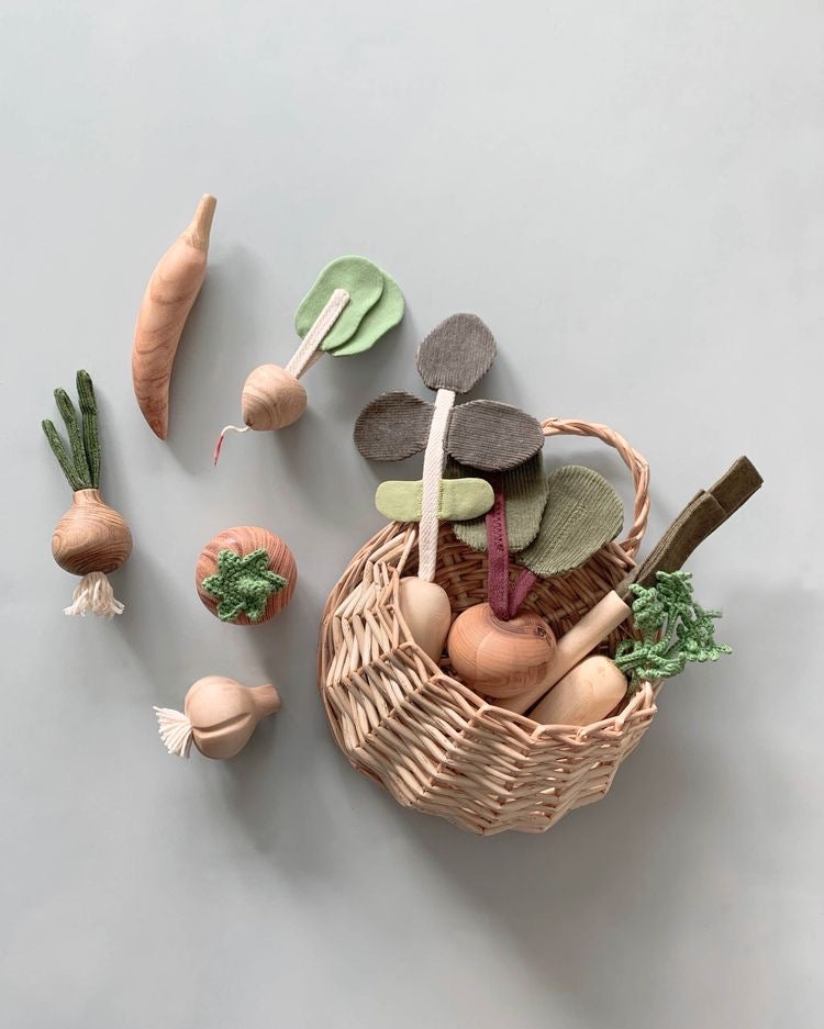 A collection of 10 Piece Handmade Wooden Vegetable Set including carrots, radishes, onions, and leeks arranged next to and inside a small wicker basket on a gray background.