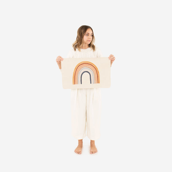 A young girl in white clothing holds up a Gathre Micro Rainbow Mat. She stands against a plain white background, looking slightly to her left, on top of a changing mat.