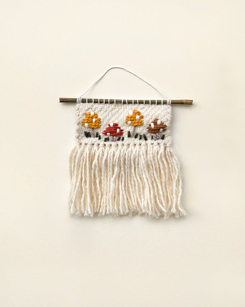 A handmade Mushroom wall hanging featuring a white fringe and a woven panel with colorful floral designs against a cream background, all suspended from a wooden dowel, perfect for nursery decor.