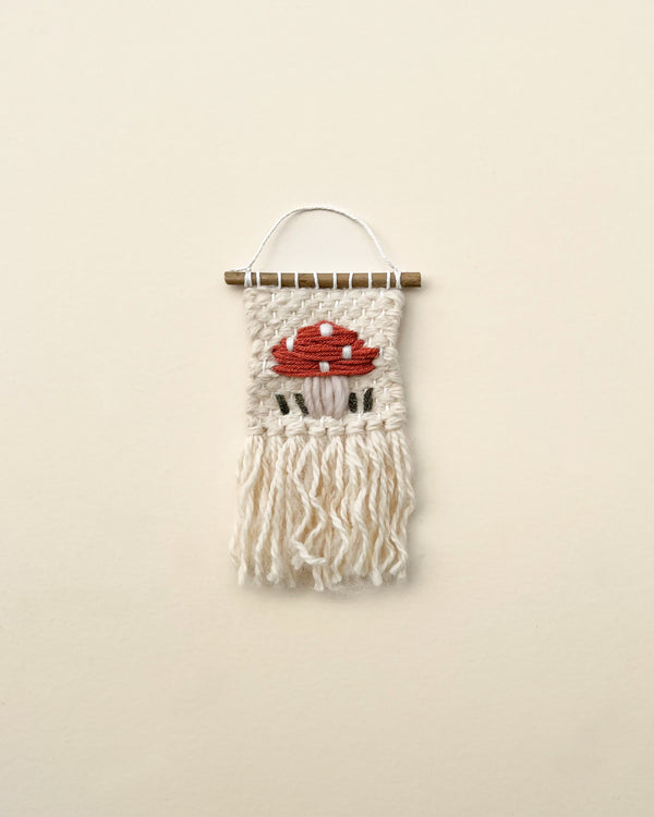 A handmade Mini Mushroom wall hanging featuring an embroidered representation of a red mushroom on a beige background, with white fringes hanging below, mounted on a horizontal stick.
